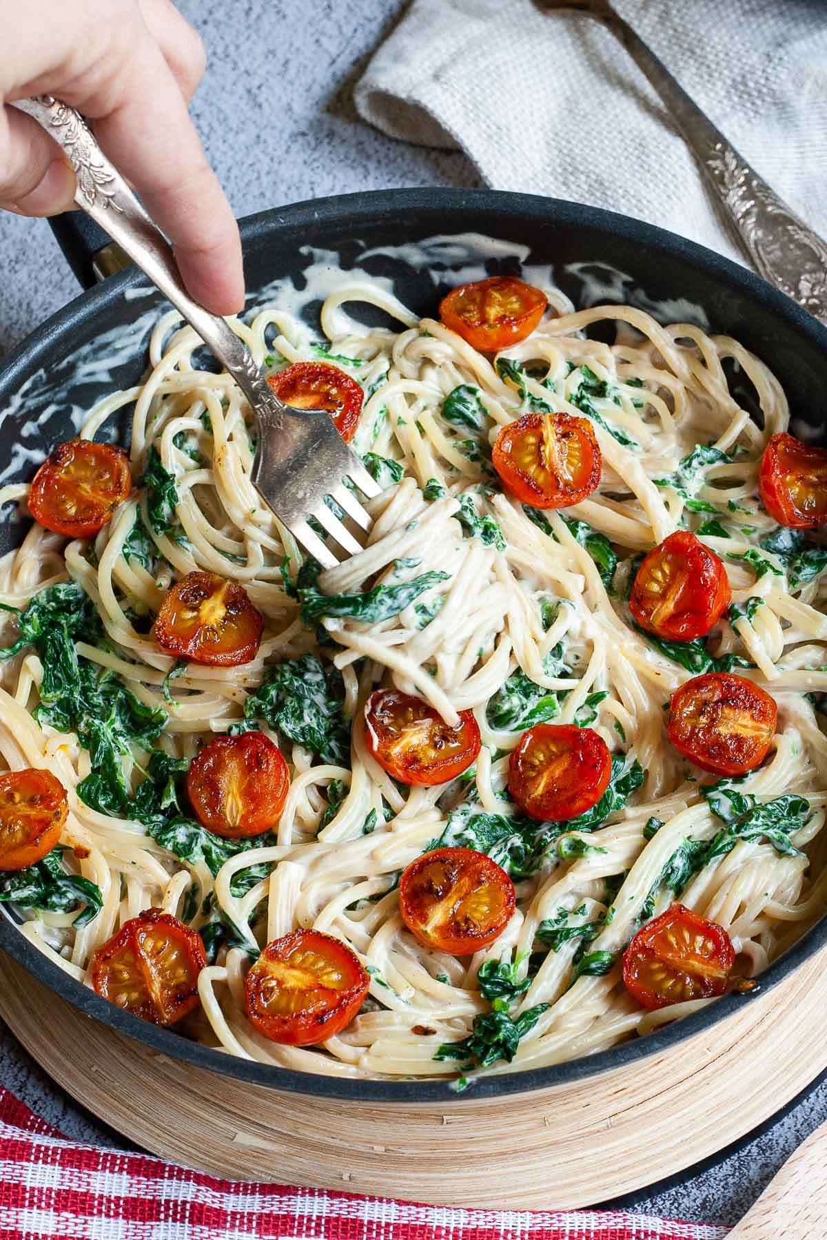 Black frying pan with spaghetti in white sauce with wilted spinach leaves and charred cherry tomatoes. A hand is holding a fork and coil up with pasta.