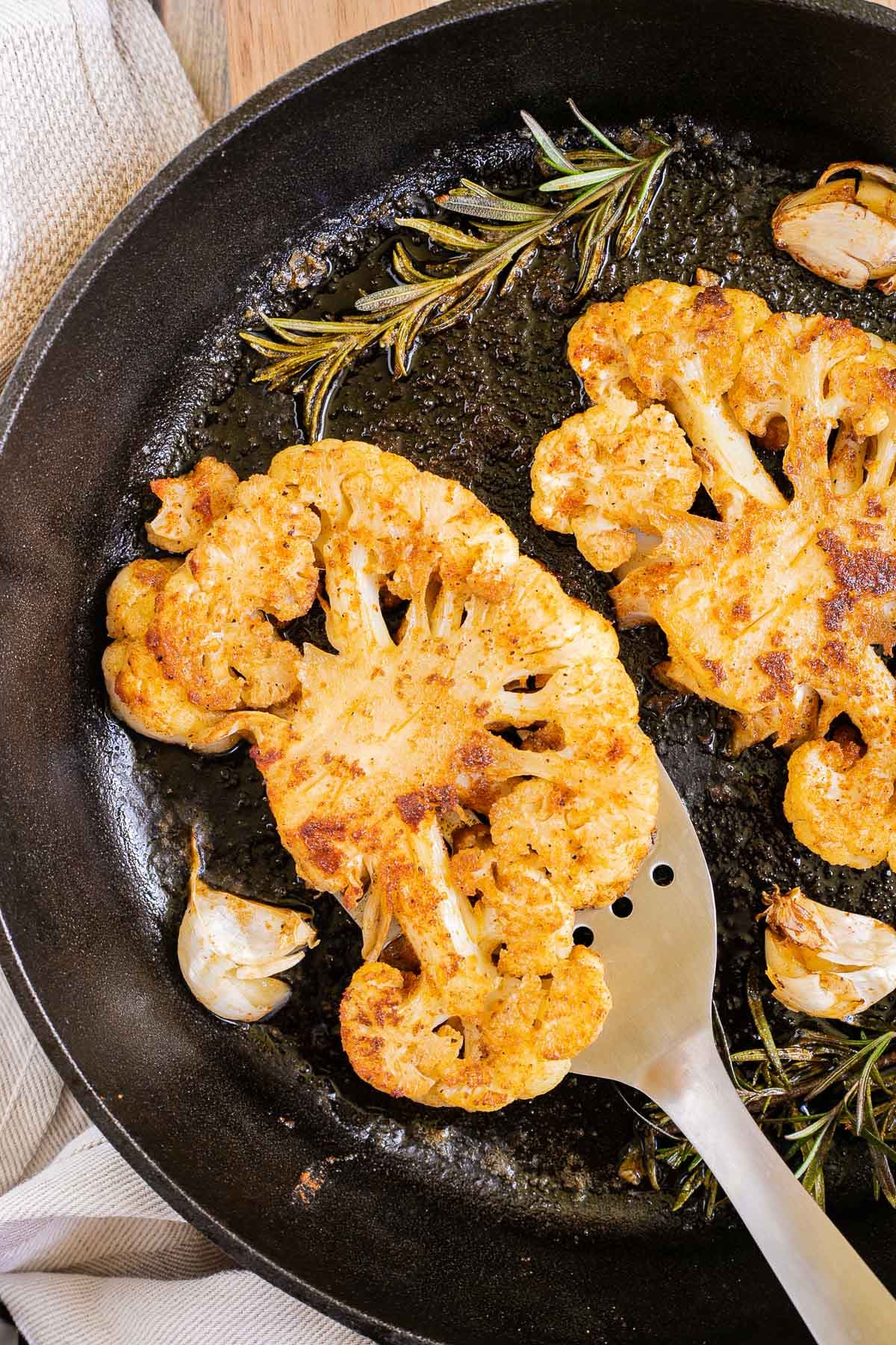 2 crispy brown yellow cauliflower slices in a cast-iron skillet surrounded by roasted garlic cloves and wilted rosemary twigs. A slotted turner is sliding underneath one.