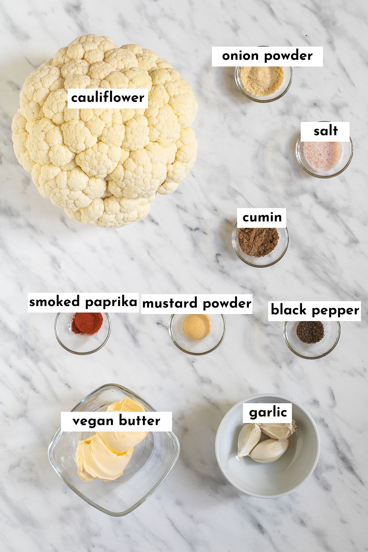 Ingredients of cauliflower steak in small glass bowls like butter, garlic, and different spices as well as a large cauliflower head next to them. 