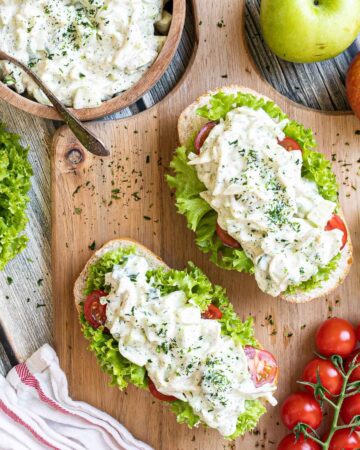 2 small loaves on a wooden board topped with lettuce leaves, cherry tomato pieces and chopped veggies in a white sauce. Leftover juicy white salad is in a wooden bowl.