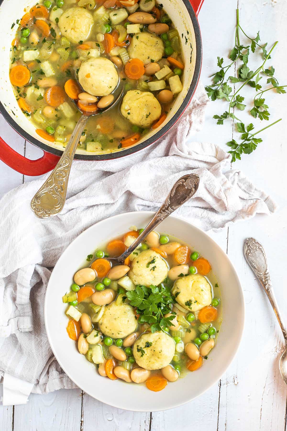 A white plate with dumplings, chopped veggies, green herbs, white beans, and green peas in a vegetable broth soup. A spoon is placed in it. The remaining soup is in a red white enameled Dutch oven next to it.
