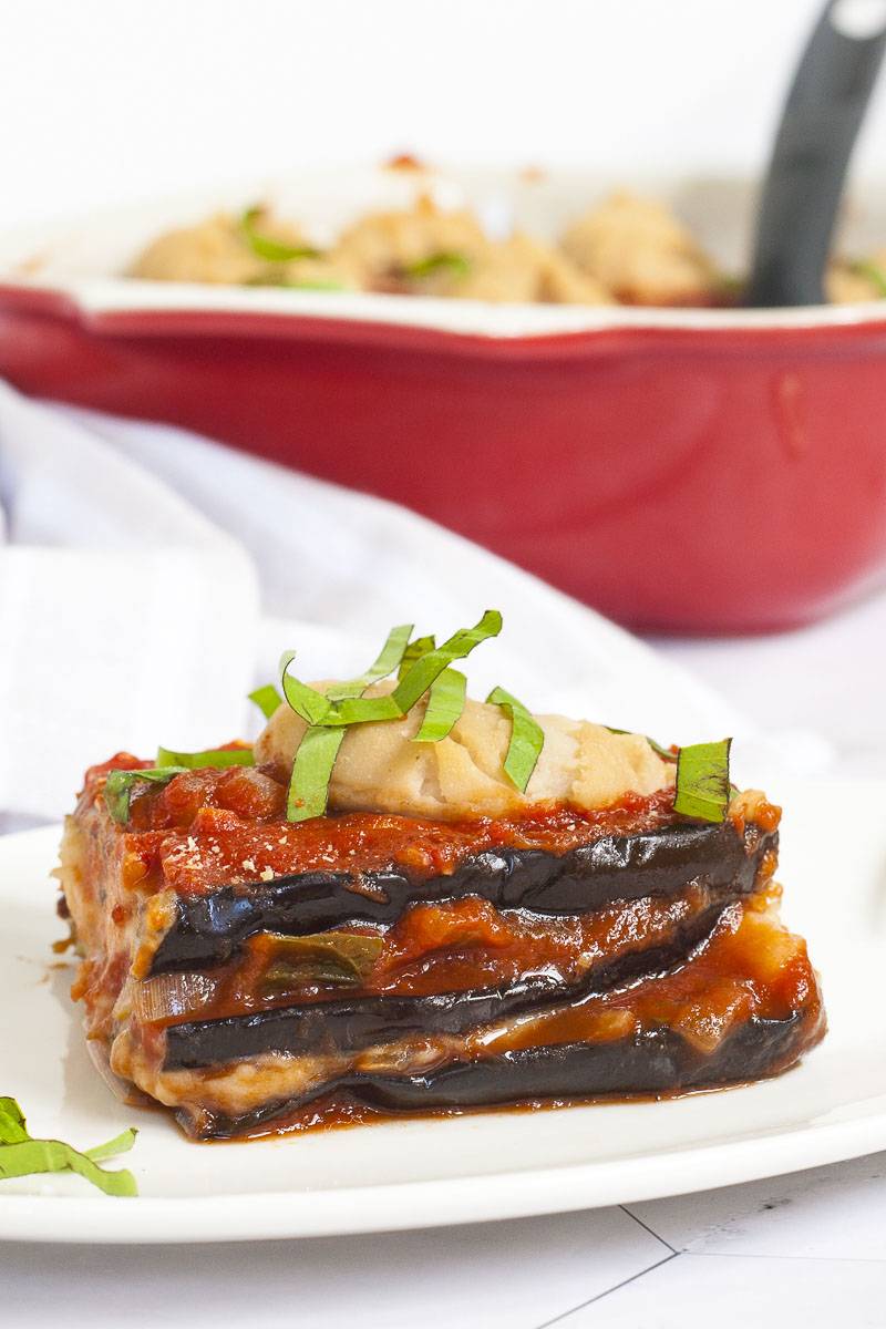 3 slices of purple eggplant in between red and white sauces on a white plate topped with fresh green herbs. In the background there is a red casserole