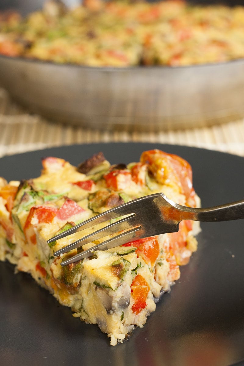 A slice of yellow frittata full of colorful vegetables on a black plate and a fork taking a bite. 