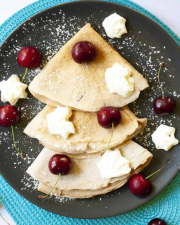 Black plate from top with 3 crepes folded as triangles with cherry and whipped cream scattered on top.