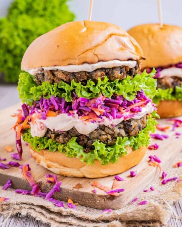 A burger bun with raffled green lettuce two lentil patties, purple coleslaw and light orange sauce.