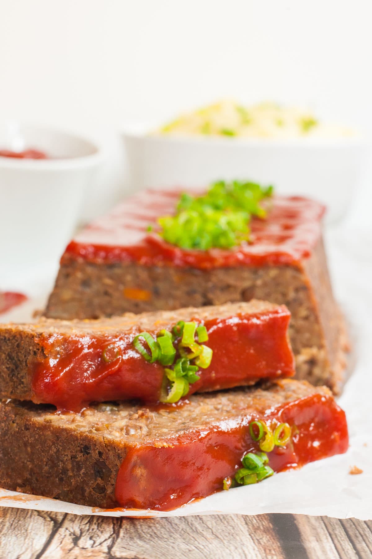 A long brown lentil loaf on parchment paper has been cut into slices. 2 slices has fallen in the front. All slices are glazed with red sauce and topped with chopped scallions. Mashed potatoes and more red glaze are placed in small white bowl in the back.