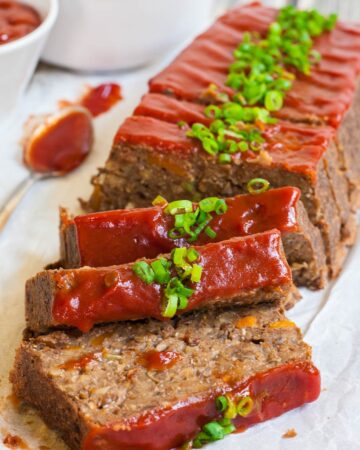 A long brown lentil loaf on parchment paper has been cut into slices. 3 slices has fallen in the front. All slices are glazed with red sauce and topped with chopped scallions.