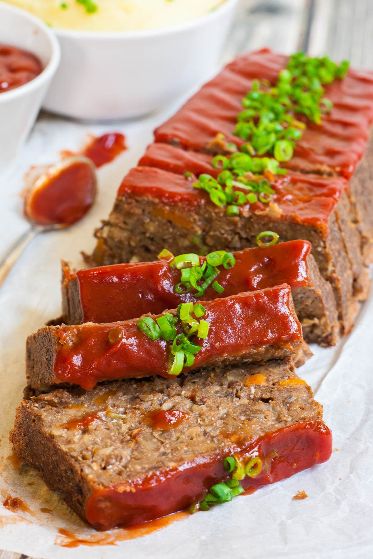 The Perfect Vegan Lentil Loaf should be deliciously rich, moist and juicy, and easy to make. Well, we give you a 10-ingredient healthy, hearty, rich and flavorful vegan lentil loaf that is not only gluten-free, but grain-free as well. #veganlentilloaf #lentiloaf #vegetarianlentilloaf #grainfree #glutenfree A long brown lentil loaf on parchment paper has been cut into slices. 3 slices has fallen in the front. All slices are glazed with red sauce and topped with chopped scallions.