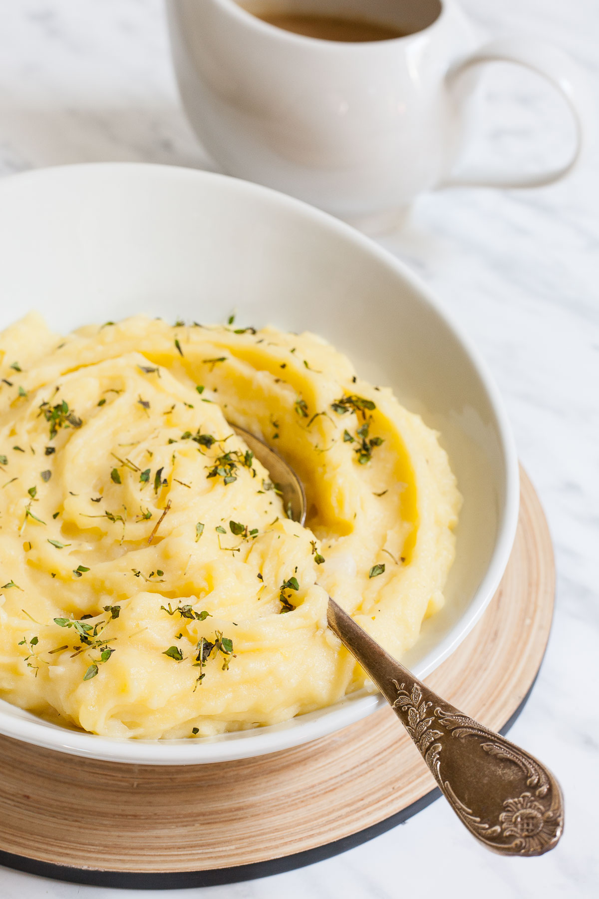 Mashed potatoes sprinkled with green herbs in a white bowl with a spoon. A small gravy boat is right behind it.