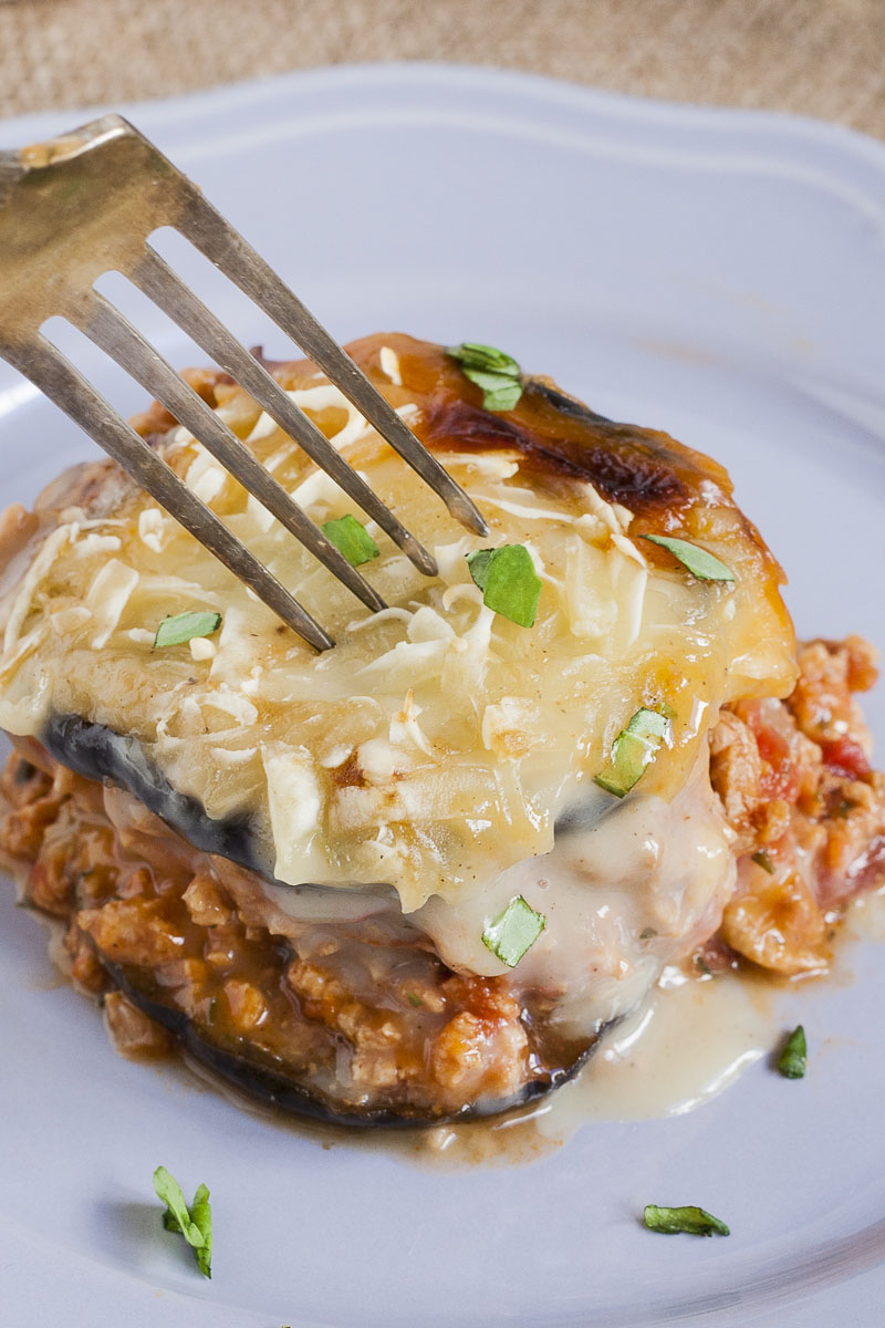 One serving of vegan moussaka is on a light purple plate with a fork. You can see the layers of purple eggplant, brown mince and white sauce on top. 