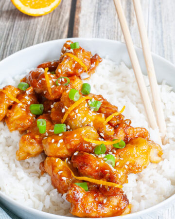 A large white bowl with white rice and sticky brown battered strips on top sprinkled with sesame seeds, orange peels and chopped spring onion