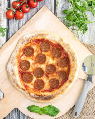 Pizza on a wooden baker's peel topped with tomato sauce, melted cheese and pepperoni slices. Fresh arugula, tomatoes and basil leaves are around it.