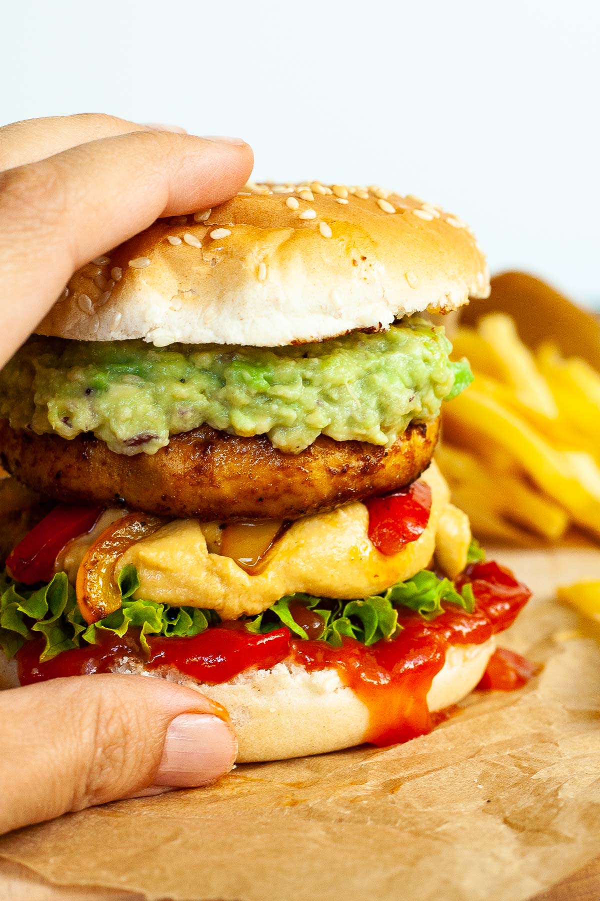 A hand is holding a burger with ketchup, lettuce, yellow cheese sauce, bell peppers, guacamole and grilled mushroom cap on brown paper. French fries are placed next to it.