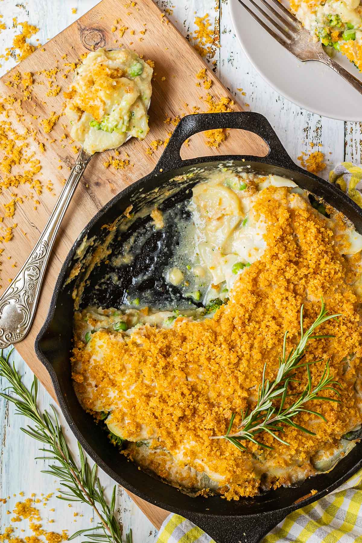 A black cast iron skillet with potato slices in a yellow thick sauce topped with yellow breadcrumbs. A part of it is missing and there is a large serving spoon next to it with some of the food from the skillet 