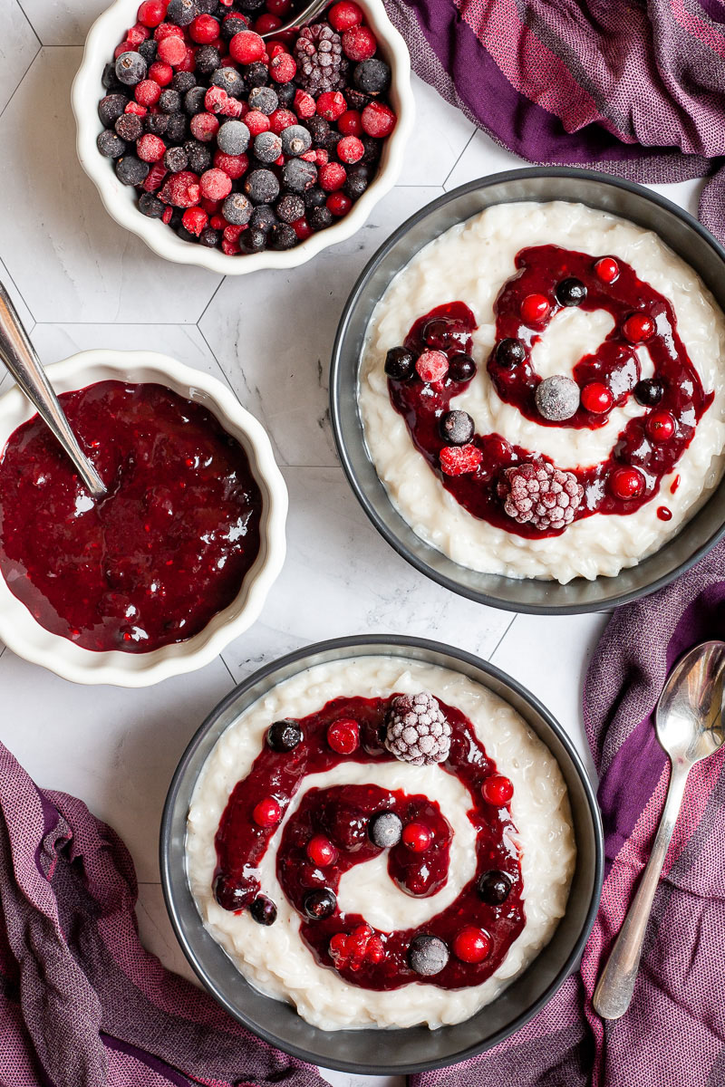 2 black bowls from above with white pudding topped with a swirl of thick red-purple sauce and different berries. 2 small white bowls next to them, one with the red sauce and one with lots of berries
