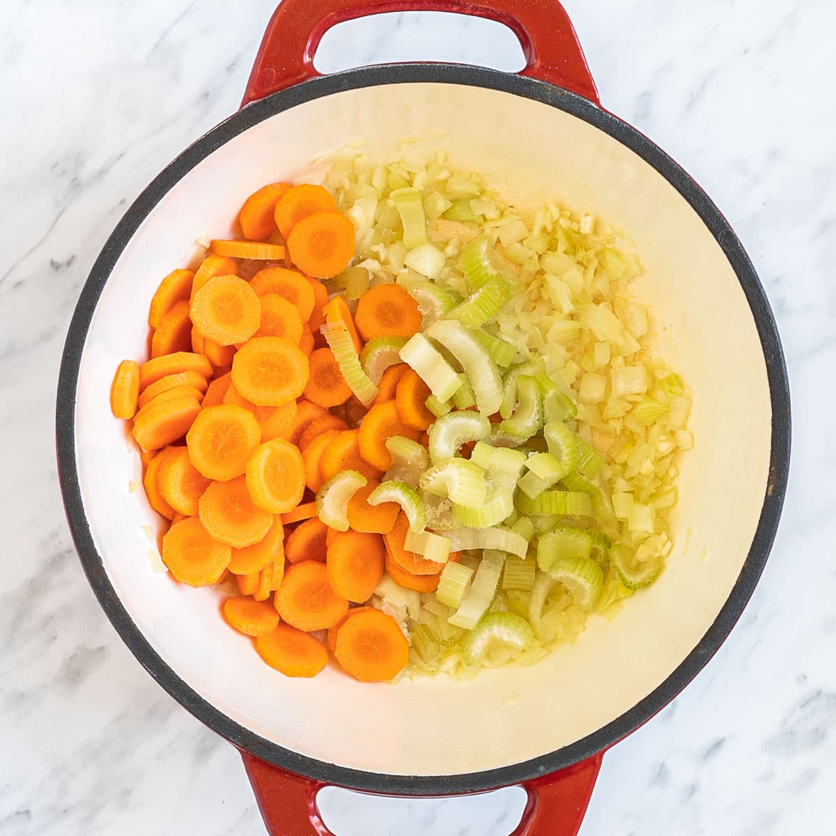 A red-white Dutch oven with chopped onion, celery, and carrot slices. 