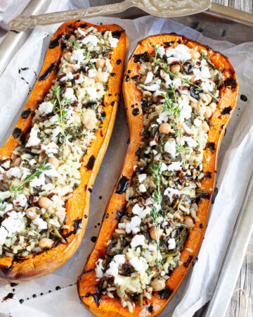 Silver baking tray from above with 2 orange butternut squash cut in half and stuffed with rice, spinach, chickpeas, olives and crumbles feta cheese drizzled with black balsamic vinegar
