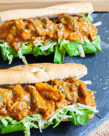 2 hoagie rolls with chopped lettuce and arugula topped with mushroom strips in a brown orange juicy sauce