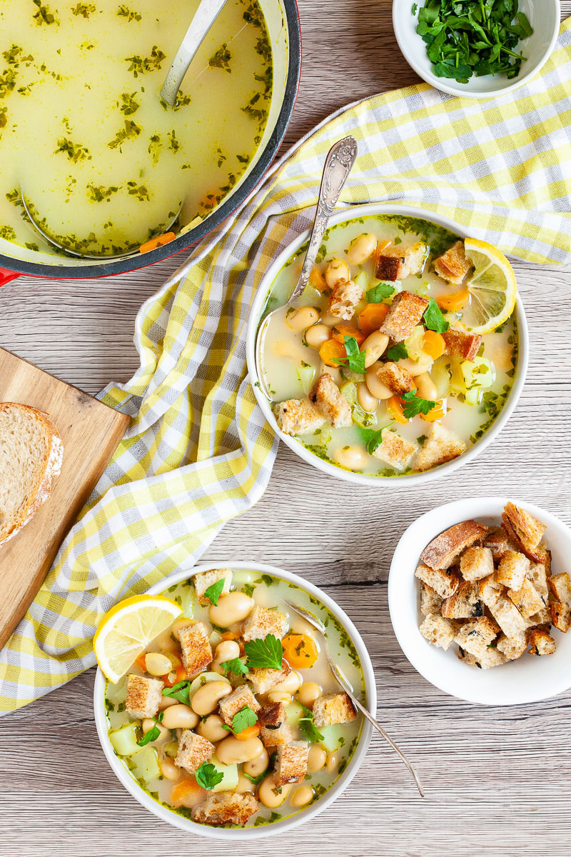 Light yellow soup served in two white bowls full of large white beans, croutons, carrot slices, diced potatoes and freshly chopped herbs. Large pot is in the corner with yellow soup. 2 small bowls with extra croutons and chopped hearbs.