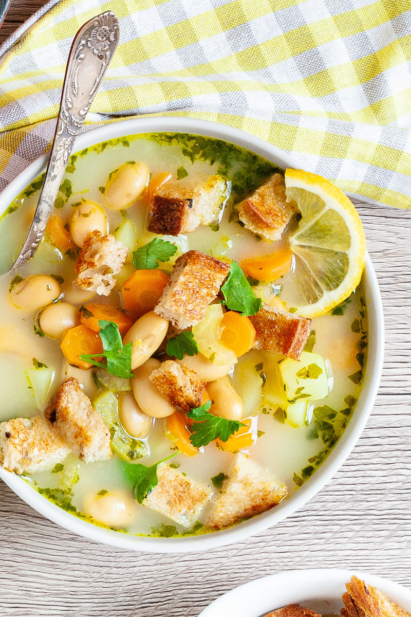 Light yellow soup served in a white bowl with a spoon full of large white beans, croutons, carrot slices, diced potatoes and freshly chopped herbs.