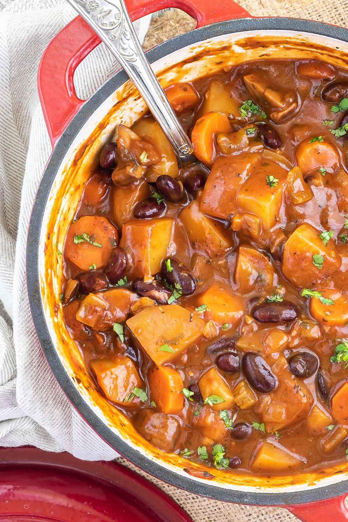 Vegetable stew in a red white enameled Dutch oven with lots of chopped vegetables like potatoes, carrots, mushroom, celery and red kidney beans. Topped with freshly chopped green herbs.