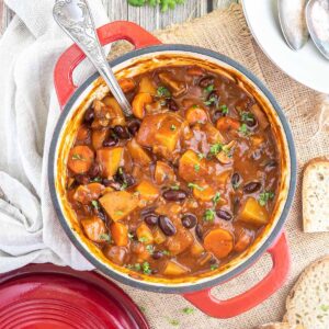 Vegetable stew in a red white enameled Dutch oven with lots of chopped vegetables like potatoes, carrots, mushroom, celery and red kidney beans. Topped with freshly chopped green herbs.
