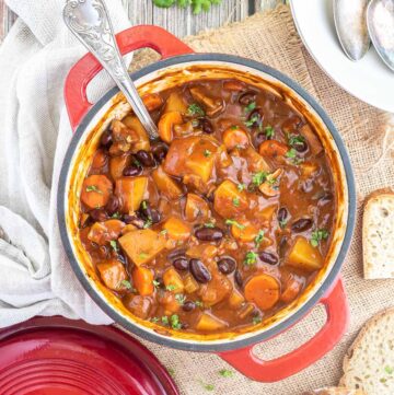 Vegetable stew in a red white enameled Dutch oven with lots of chopped vegetables like potatoes, carrots, mushroom, celery and red kidney beans. Topped with freshly chopped green herbs.