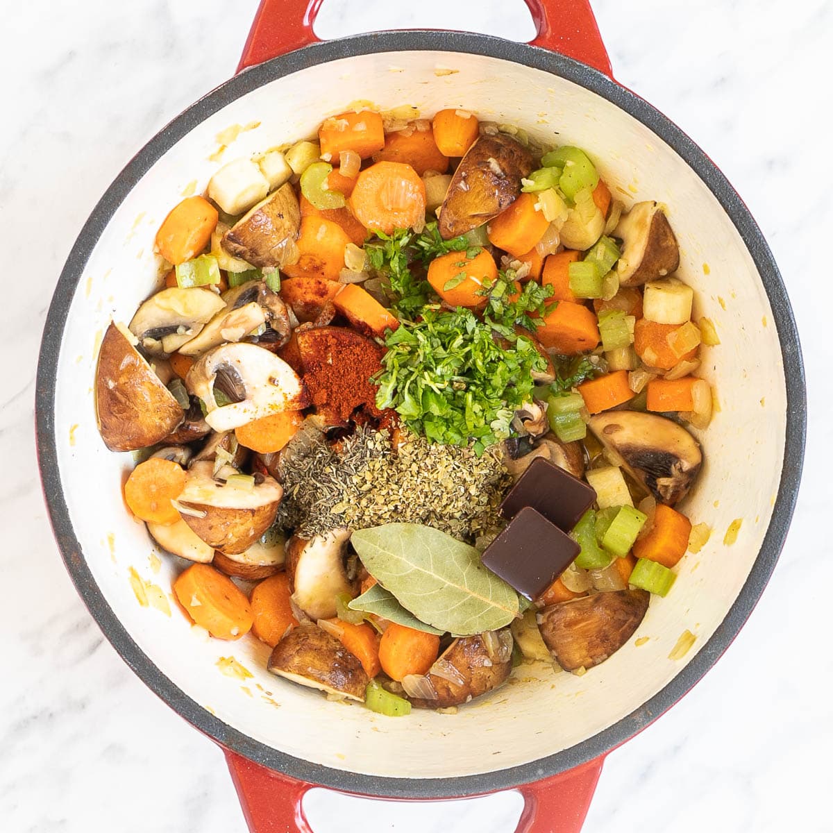 Red white enameled Dutch oven with lots of chopped veggies like carrots, mushrooms, celery and small heaps of green herbs and red spices