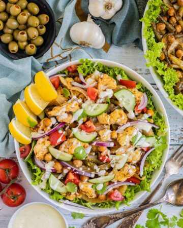 A large white bowl with green salad leaves topped with cauliflower florets, green beans, chickpeas, fresh tomatoes drizzled with a white sauce. Olives and more roasted veggies are next to it on small plates.