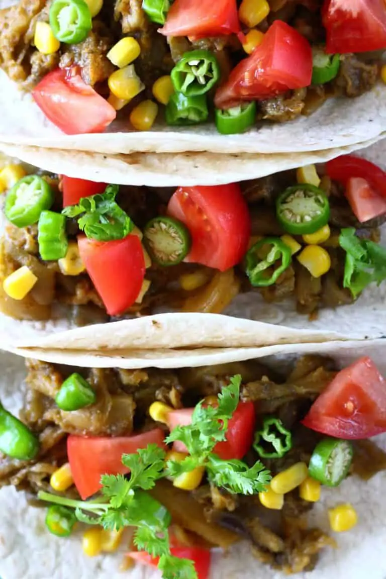 Photo of three tacos on a blue plate filled with roasted eggplant, sweetcorn, sliced green chilli, chopped tomatoes.
