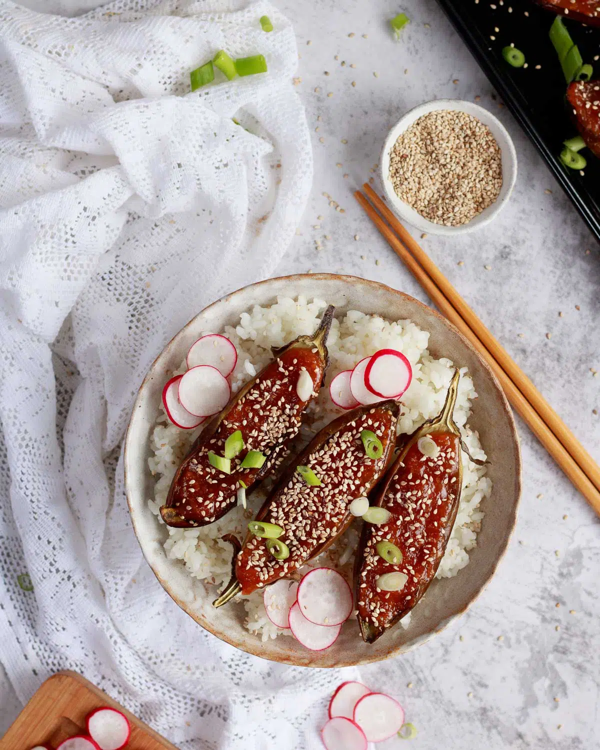 A bowl of japanese rice with miso aubergine laid on top. There are sesame seeds, radish and spring onions to garnish. 
